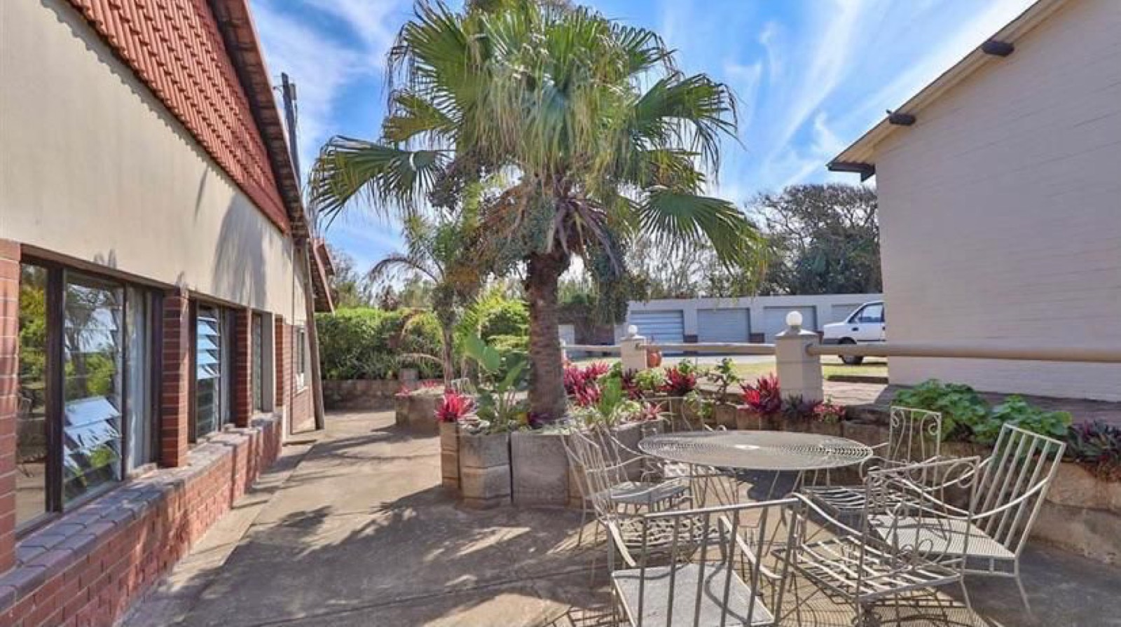 Tetelestai Recovery Centre courtyard patio with a tall palm tree, wrought-iron outdoor table and chairs, and flowering garden beds